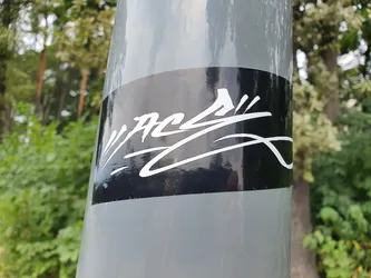 A black and white sticker with a graffiti-style design on a gray pole. The sticker has a glossy finish and the design is in white against a black background. The background shows green vegetation, indicating an outdoor setting.