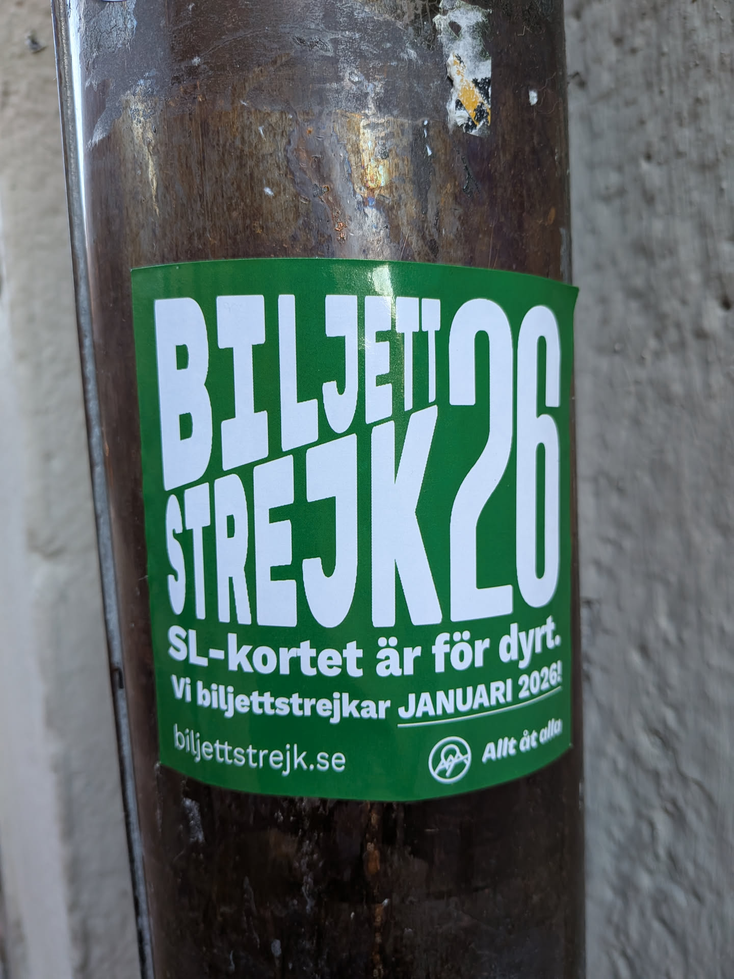 A rectangular sticker with a vibrant green background and bold white text. The prominent headline reads 'BILJETTSTREJK26'. Below this, smaller white text states 'SL-kortet är för dyrt. Vi biljettstrejkar JANUARI 2026!'. At the bottom, a website address 'biljettstrejk.se' is displayed alongside a circular logo featuring a stylized 'A' and the phrase 'Allt åt alla'. The sticker is affixed to a dark, weathered, cylindrical pole.