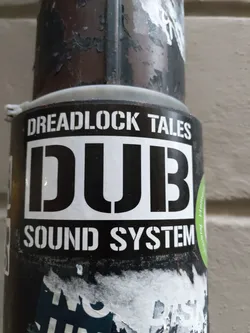 Street sticker A black and white sticker with the text "Dreadlock Tales" at the top and "DUB Sound System" at the bottom, with the word "DUB" prominently displayed in a large, bold font in the center. The sticker is affixed to a weathered, dark-colored metal surface.