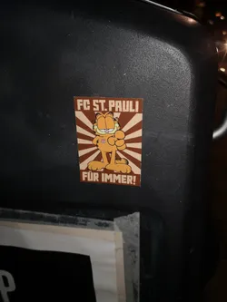 Street sticker The sticker prominently features the iconic orange cartoon cat, Garfield, standing confidently with one hand on his hip and the other clenched in a fist, giving a thumbs-up. He sports a small, circular FC St. Pauli logo on his chest. The background is composed of radiating stripes in alternating shades of brown and light beige, creating a sunburst effect. Bold, blocky text in brown reads "FC ST. PAULI" at the top and "FÜR IMMER!" (meaning "FOREVER!") at the bottom.