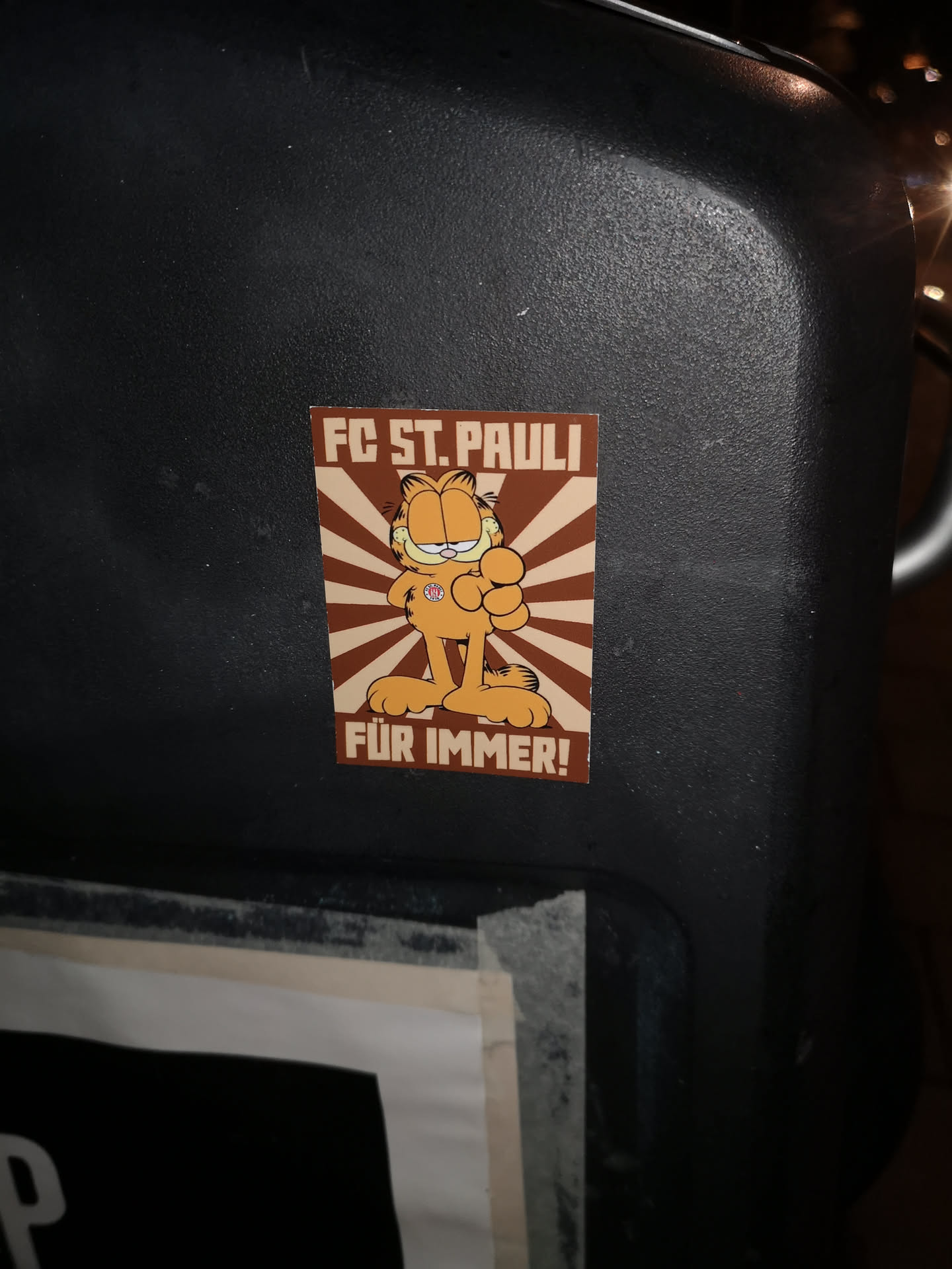The sticker prominently features the iconic orange cartoon cat, Garfield, standing confidently with one hand on his hip and the other clenched in a fist, giving a thumbs-up. He sports a small, circular FC St. Pauli logo on his chest. The background is composed of radiating stripes in alternating shades of brown and light beige, creating a sunburst effect. Bold, blocky text in brown reads "FC ST. PAULI" at the top and "FÜR IMMER!" (meaning "FOREVER!") at the bottom.