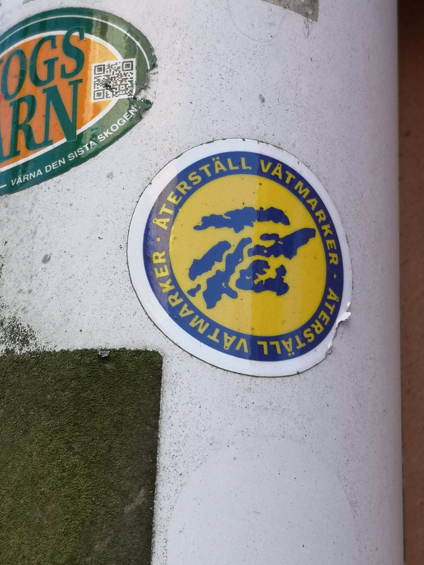 A circular sticker with a bright yellow background at its core, featuring an irregular, organic blue shape in the center that resembles a body of water or a wetland viewed from above. This central design is encircled by a dark blue ring, which prominently displays the white capitalized text 'ÅTERSTÄLL VÅTMARKER' repeated twice, separated by small white dots. The entire sticker is framed by a thin white outer border. The sticker shows minor signs of wear, including a slight peel on its bottom right edge.