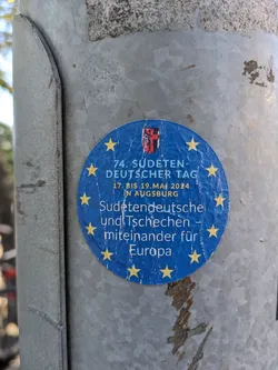 A blue circular sticker commemorating the 74th Sudeten German Day, held in Augsburg from May 17th to 19th, 2024. The sticker features yellow stars and text in German, emphasizing unity between Sudeten Germans and Czechs for Europe.