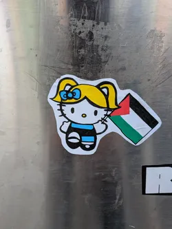 Street sticker The sticker features a cartoon character reminiscent of Hello Kitty, but with distinct human-like features including yellow hair styled in pigtails and a blue bow. The character has a white face, black oval eyes, and a small yellow oval nose. It is depicted wearing a black and blue striped outfit. The character is holding a small, rectangular Palestinian flag, which is colored red, white, green, and black. The sticker has a visible white border, indicating it was cut out, and is affixed to a scratched, metallic surface.