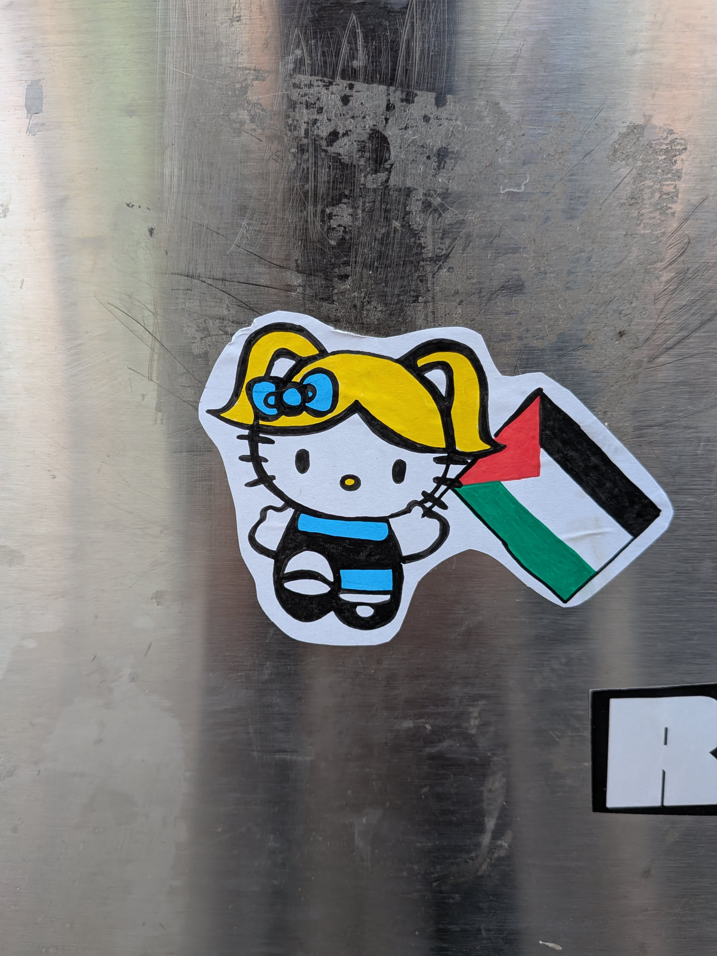 The sticker features a cartoon character reminiscent of Hello Kitty, but with distinct human-like features including yellow hair styled in pigtails and a blue bow. The character has a white face, black oval eyes, and a small yellow oval nose. It is depicted wearing a black and blue striped outfit. The character is holding a small, rectangular Palestinian flag, which is colored red, white, green, and black. The sticker has a visible white border, indicating it was cut out, and is affixed to a scratched, metallic surface.