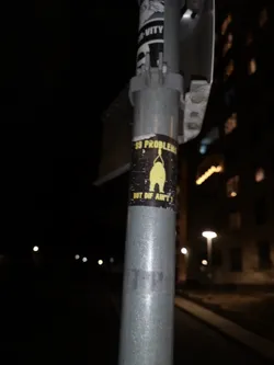 Street sticker A rectangular sticker featuring a stylized, somewhat stout yellow figure lifting a barbell or heavy object above its head, set against a black background. The figure has a simple, almost cartoonish design. Above the figure, in yellow capital letters, is the text '99 PROBLEMS'. Below the figure, also in yellow capital letters, is 'BUT DIS AIN'T 1!'. The sticker shows signs of wear and tear, with some white scuffs and faded areas, particularly around the edges and within the black background, indicating it's a physical sticker on a metal pole.