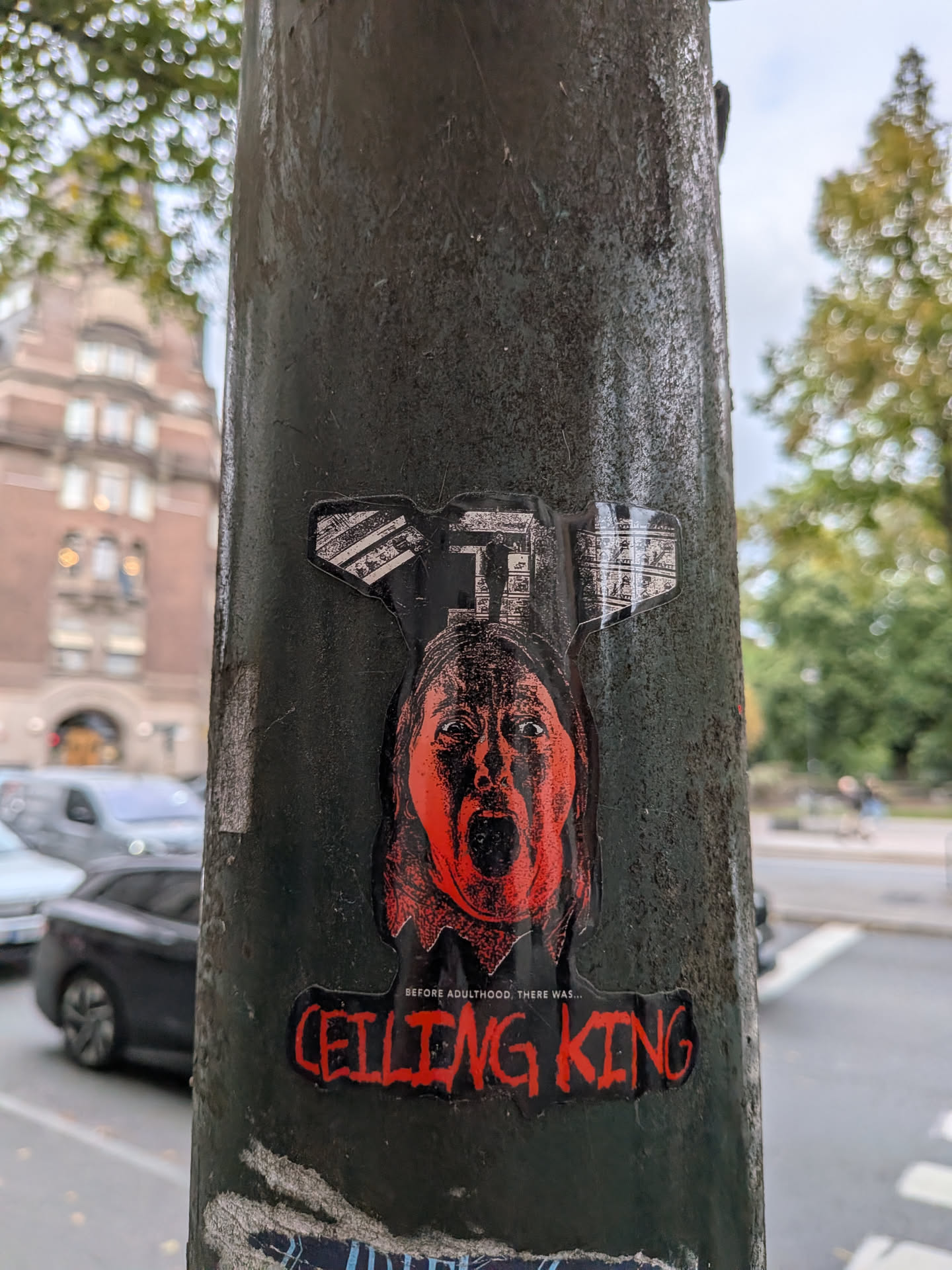 A die-cut sticker affixed to a weathered metal pole. The central image is a high-contrast, red and black illustration of a person screaming in terror. Above the head is a black and white graphic depicting a ceiling fan. Below the face, small white text reads 'BEFORE ADULTHOOD, THERE WAS...', followed by the main title 'CEILING KING' in a bold, dripping red horror-style font.