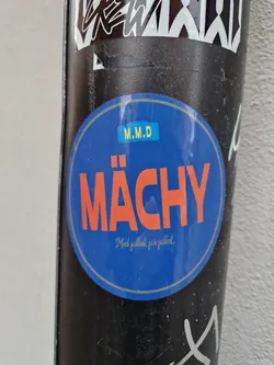 Street sticker A round blue sticker with the word "Mächy" in large orange letters.  Smaller text below reads "Med pallett, för pallett." (With pallet, for pallet).  There's a small yellow rectangle with "M.M.D" inside at the top of the sticker.