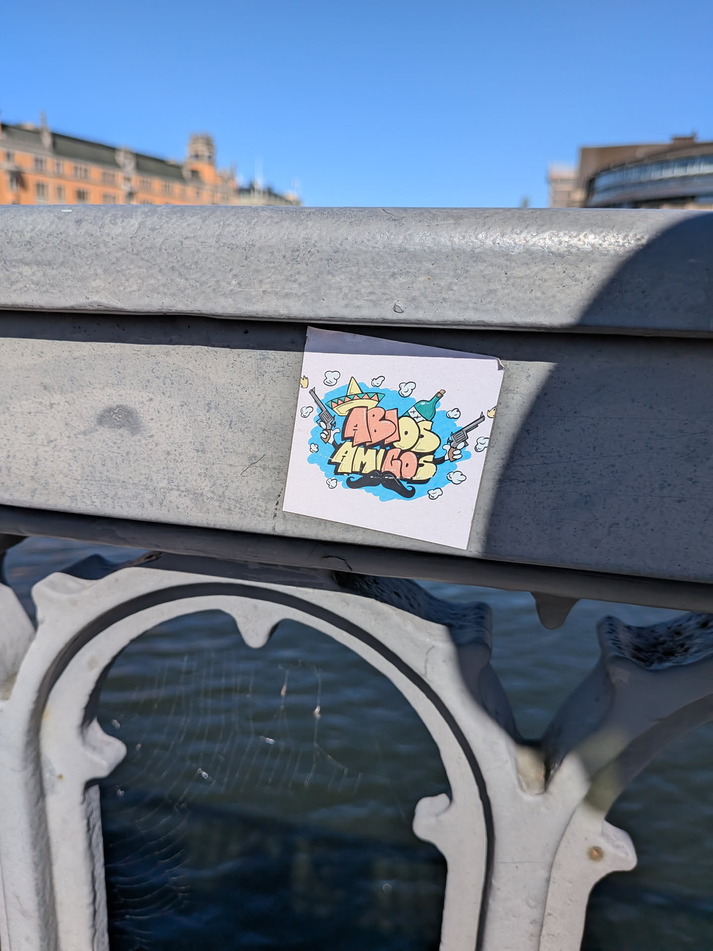 The sticker is square-shaped, featuring a vibrant, cartoonish design on a white background. The central element is the text 'ABDOS AMIGOS' in large, bubble-style, graffiti-like letters. 'ABDOS' is colored in shades of orange and red, while 'AMIGOS' is in yellow and green. Surrounding the text are various elements: a yellow sombrero with a red band on the top left, a green bottle (resembling tequila) with an 'X' on it and a silver revolver with a brown handle firing a bullet on the right, and a prominent black, stylized mustache at the bottom. Light blue cloud-like shapes or smoke puffs are scattered around the text and elements, adding to the dynamic feel. The overall aesthetic is playful, energetic, and festive.