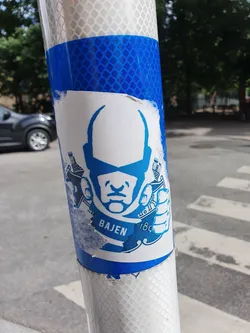 A blue and white sticker featuring a stylized face and the text "Bajen 1897".  The sticker is affixed to a reflective blue pole.