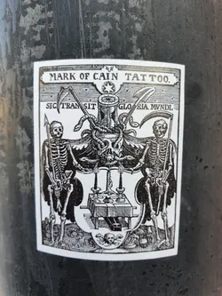 Street sticker Black and white sticker with a vintage woodcut style illustration featuring two skeletons, scythes, snakes, an hourglass, and a skull. The text at the top reads "MARK OF CAIN TATTOO."  Latin text is visible below the top text: "SIC TRANSIT GLORIA MUNDI". The sticker appears to be advertising a tattoo parlor.