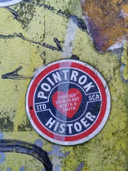 A round sticker with a red and white border and a dark blue center. The text on the sticker says "POIN TROK HIS TOER" and contains a heart with the text "THROUGH YOUR HEART NORTH & SOUTH".  The abbreviation ITD and SCA are also visible.