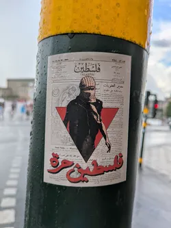 Street sticker The sticker, designed to resemble an old newspaper clipping, features a central figure wearing a keffiyeh and sunglasses, with a determined expression. This figure is partially obscured by a prominent red inverted triangle, a symbol often associated with resistance. Behind the figure, faint grayscale illustrations depict historical or symbolic scenes, including a crescent moon and star, and what appears to be a building or landscape. The background is filled with small, dense Arabic text, mimicking newspaper articles. Large, stylized red Arabic text at the top reads 'فلسطين' (Falastin) and 'FALASTIN' in English, while equally prominent red Arabic text at the bottom declares 'فلسطين حرة' (Falastin Hurra), meaning 'Free Palestine'. The sticker is adhered to a dark green pole, which is visibly wet with raindrops, adding to its authentic street art appearance.