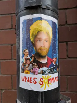 Street sticker The sticker is a collage featuring a large portrait of a man with blond hair and a beard against a blue, striped background. Below him, a collection of smaller images depict various people and a vintage camper van. The people include a woman in a blue dress, a man in a striped shirt, a child in a yellow shirt, another child in a blue and white striped shirt, a woman in pink, and another man with long hair. The camper van has people inside. The bottom of the sticker has the text "UNES SOMMAR" in red, with a stylized yellow sun replacing the 'O' in 'SOMMAR'. The sticker has a white border and appears to be slightly creased.
