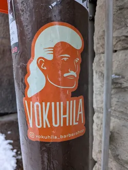 Street sticker The sticker features a stylized profile illustration of a man's head, facing right, rendered in white against a vibrant orange background. He sports a distinctive mullet hairstyle, with shorter hair on top and longer, wavy hair at the back, and a prominent mustache. Below the illustration, the word "VOKUHILA" is written in large, bold white capital letters. Further below, there's a small white Instagram logo followed by the handle "vokuhila_barbershop" in white text. The sticker has an irregular, rounded shape that follows the contours of the design, and it is affixed to a dark brown, textured pole.