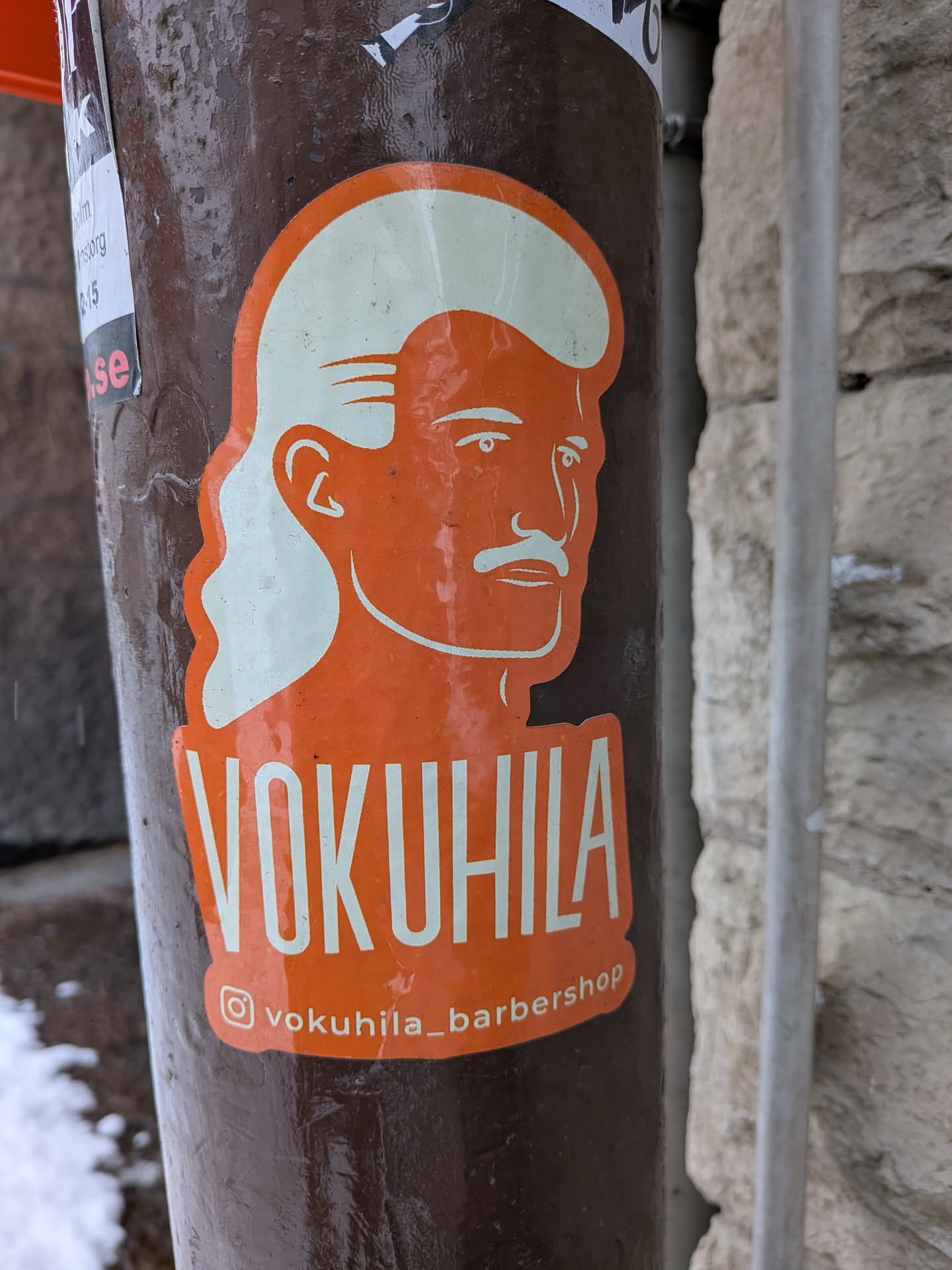 The sticker features a stylized profile illustration of a man's head, facing right, rendered in white against a vibrant orange background. He sports a distinctive mullet hairstyle, with shorter hair on top and longer, wavy hair at the back, and a prominent mustache. Below the illustration, the word "VOKUHILA" is written in large, bold white capital letters. Further below, there's a small white Instagram logo followed by the handle "vokuhila_barbershop" in white text. The sticker has an irregular, rounded shape that follows the contours of the design, and it is affixed to a dark brown, textured pole.