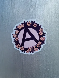 Street sticker Round sticker with a pink anarchy symbol in the center, surrounded by a wreath of peach-colored flowers and dark purple leaves. The sticker is on a silver surface.