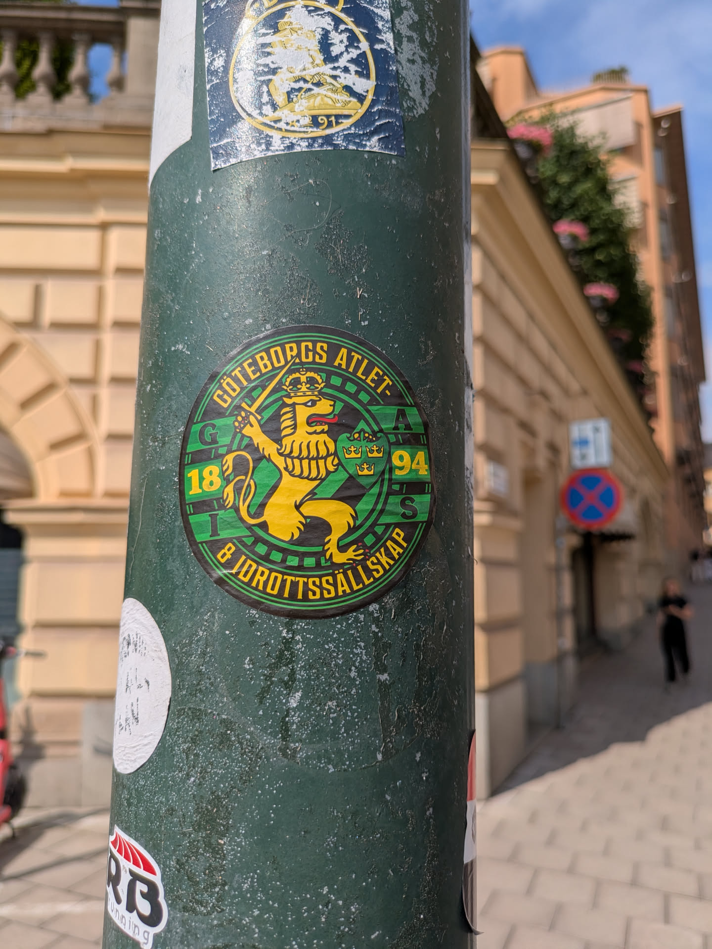Street sticker The sticker is circular, featuring a vibrant yellow crowned lion rampant holding a sword, set against a checkered green and black background. The lion has a visible red tongue. An outer black ring frames the central design, containing yellow text. Inside the main emblem, the letters 'G', 'A', and 'S' are positioned around the lion, along with the numbers '18' and '94'. The overall design evokes a classic sports or athletic club crest.