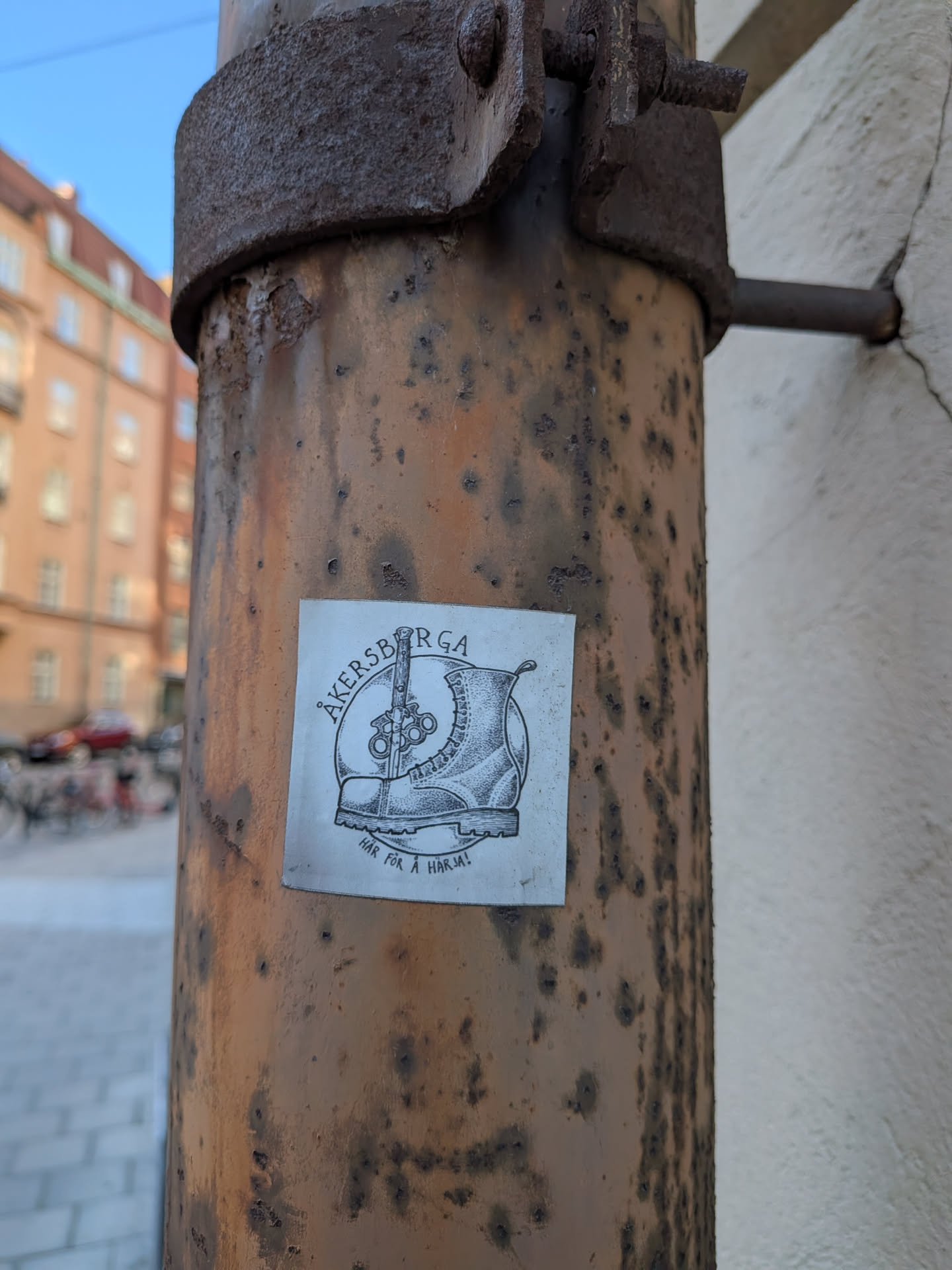 The sticker is a square, black and white illustration adhered to a rusty metal pole. It features a detailed combat boot on the right, with laces and stitching visible. Behind the boot, a large, stylized knife points upwards. To the left of the boot, a pair of brass knuckles is depicted. The entire central image is enclosed within a circular border. Above the circle, arched text reads "ÅKERSBERGA". Below the main illustration, smaller text reads "HÄR FÖR Å HÄRJA!". The sticker shows signs of wear and slight discoloration, consistent with outdoor exposure.