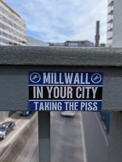 Street sticker A rectangular sticker featuring a bold, three-tiered design. The top and bottom sections are blue, while the middle section is black. The top blue section displays 'MILLWALL' in large white capital letters, flanked by two circular Millwall Football Club logos. Each logo shows a white lion rampant within a circle, with 'MILLWALL FOOTBALL CLUB' and '1885' inscribed around it. The central black section reads 'IN YOUR CITY' in prominent white capital letters. The bottom blue section completes the message with 'TAKING THE PISS' in white capital letters. The overall aesthetic is strong and assertive.