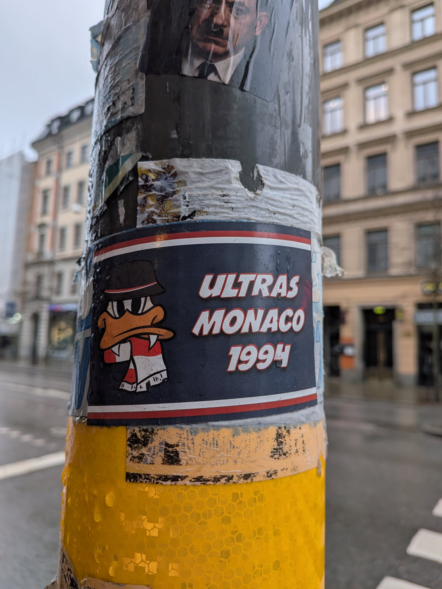 The sticker is rectangular with a dark blue background, framed by thin red and white stripes. On the left, a cartoon duck character is depicted with an orange beak, white eyes with black pupils, wearing a black bucket hat with a red and white stripe, and a red and white striped scarf. The duck has a somewhat determined or aggressive expression. To the right, in bold white capital letters, are the words "ULTRAS MONACO" stacked above "1994". The sticker shows some wear and water droplets, indicating its outdoor placement on a pole.