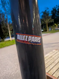 Street sticker A rectangular sticker adhered to a dark, reflective pole. The sticker features a dark blue background with a thin red and yellow border. The main text reads 'ALLEZ PARIS' in bold, white, stylized capital letters with a black outline. Below the text, there are detailed illustrations of two hand flares lying horizontally: a red one on the left and a blue one on the right, both with the small text 'HAND FLARE' printed on them.