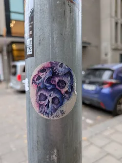Street sticker The sticker is circular and features a detailed illustration of multiple skulls intertwined. The skulls are depicted in shades of deep blue, purple, and pink, with intricate details in their eye sockets and teeth. The overall style is somewhat gothic and macabre. There is some handwritten text and numbers in the lower right corner of the sticker.