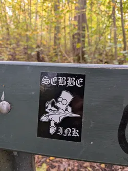 Street sticker The sticker is predominantly black with white graphics. It features a stylized depiction of Bart Simpson from The Simpsons, wearing glasses and drinking from a cup with a straw. His body appears to be melting or dissolving into a liquid form. Above Bart's head, in a gothic-style font, is the text 'SEBBE'. Below Bart's feet, also in a gothic-style font, is the text 'INK'. The overall design has a slightly edgy or rebellious feel.