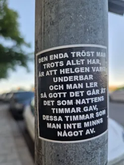 Street sticker This sticker features a passage of text, apparently in Swedish, and it seems to reflect some amount of light, giving it a metallic sheen. The sticker is stuck to a grey metal pole. The text on the sticker reads: "DEN ENDA TRÖST MAN TROTS ALLT HAR, ÄR ATT HELGEN VARIT UNDERBAR OCH MAN LER SÅ GOTT DET GÅR ÅT DET SOM NATTENS TIMMAR GAV, DESSA TIMMAR SOM MAN INTE MINNS NÅGOT AV."