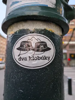 Street sticker The sticker is oval-shaped with a white background and a black border. It features two black bucket hats with white lettering. The left hat has an 'S' and the right hat has an 'M'. Behind the hats, there are stylized black outlines of mountains. Below the hats, in black, is the text 'dva klobúky'. The sticker appears to be slightly worn but clearly visible.