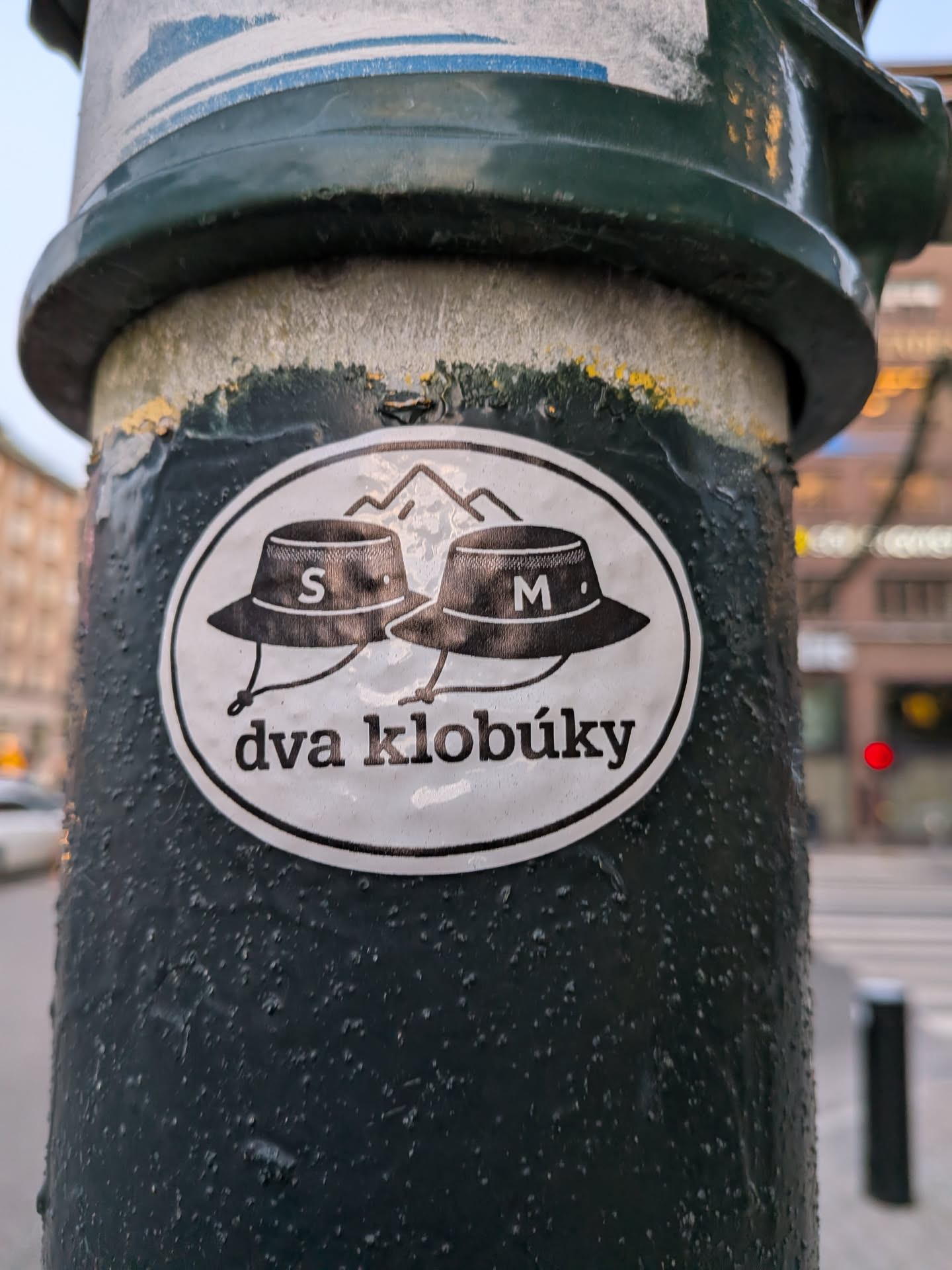 The sticker is oval-shaped with a white background and a black border. It features two black bucket hats with white lettering. The left hat has an 'S' and the right hat has an 'M'. Behind the hats, there are stylized black outlines of mountains. Below the hats, in black, is the text 'dva klobúky'. The sticker appears to be slightly worn but clearly visible.