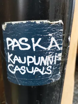A dark blue sticker with white graffiti-style text reading 'Paska Kaupunki Casuals'. The sticker is affixed to a dark pole and shows some signs of wear and tear.