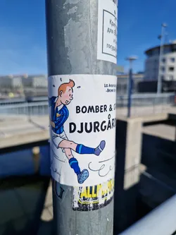 Street sticker A sticker featuring a cartoon illustration of a soccer player, possibly inspired by Tintin, with the text "Bomber & Djurgården."  The sticker is affixed to a metal pole outdoors.