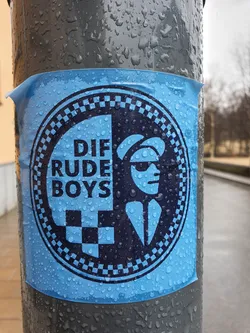 Street sticker A blue circular sticker featuring the logo for the band Die Rude Boys. The logo includes a checkered pattern, a stylized image of a man in a suit, and the band's name. The sticker is affixed to a wet pole.