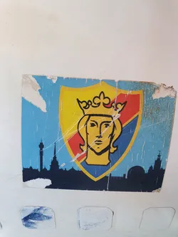 Street sticker A weathered sticker depicting a stylized crowned figure on a shield, set against a backdrop of the Riga skyline. The sticker shows signs of age and wear.