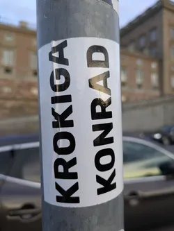 Street sticker The sticker is predominantly white with bold, black sans-serif lettering. The text is oriented vertically, with 'KROKIGA' on the left and 'KONRAD' on the right. The sticker is wrapped around a cylindrical, metallic pole, and the edges of the sticker appear slightly worn or creased, suggesting it has been applied for some time. The background is blurred, showing a building and a car.