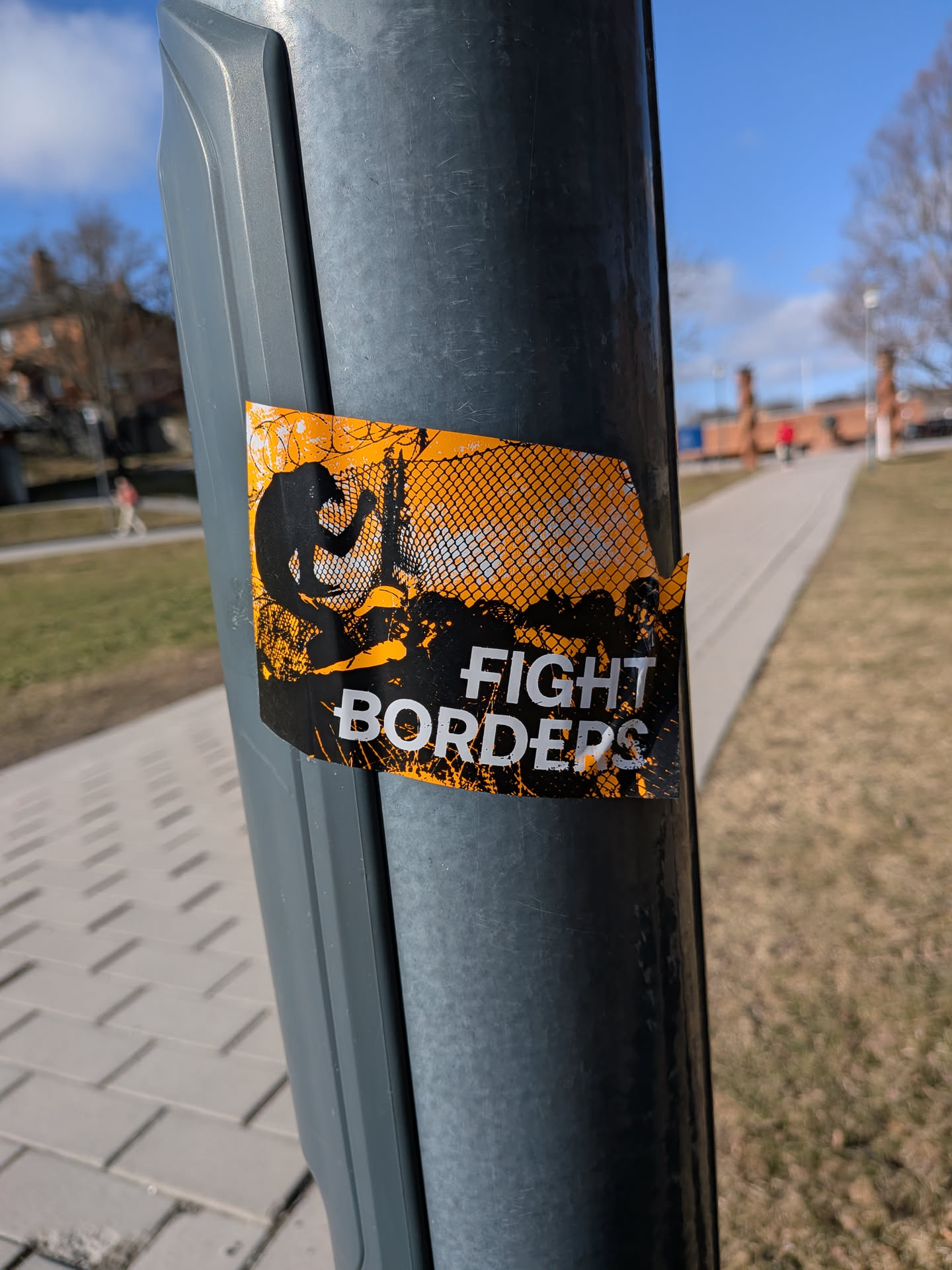 The sticker is rectangular with intentionally torn or distressed edges, giving it a rugged appearance. Its primary colors are a bright, almost neon orange and stark black, with white text. The background is predominantly orange, overlaid with a black, grungy, and splattered texture that suggests dirt or struggle. On the left side, a black silhouette of a person is depicted in the act of climbing over a fence or barrier, which is rendered with a mesh pattern and barbed wire elements in black against the orange. The right side of the sticker features bold, white, sans-serif text that reads "FIGHT BORDERS", with "FIGHT" positioned above "BORDERS". The overall design is high-contrast and impactful, conveying a strong message.