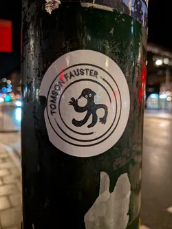 Street sticker A circular white sticker featuring a central, black, cartoonish, multi-limbed creature. The creature has a small head with two large white eyes (black pupils), a beak-like mouth, and what appears to be a small cap or hat. Its body is amorphous with several tentacle-like limbs, one of which seems to be holding a small stick or tool. The text "TOMSON FAUSTER" is written in black, sans-serif capital letters along the top inner edge of the circle, framed by two thin black concentric lines. The sticker is applied to a dark, worn, and scratched pole, with reflections of city lights visible in the background.