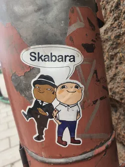 A sticker depicting two cartoon characters. One is wearing a suit and hat, and the other is wearing a polo shirt and jeans. A speech bubble above them says "Skabara". The sticker is applied to a weathered metal pole.