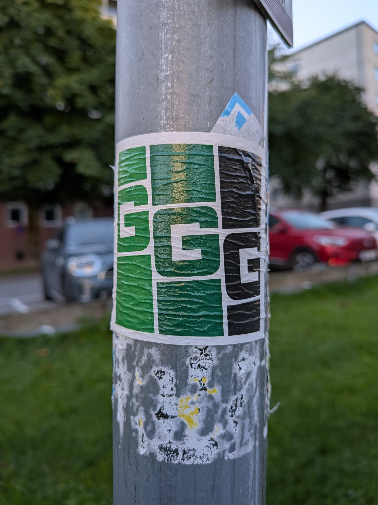The sticker is predominantly green and black, with white outlines separating sections. It features a grid-like pattern where each cell contains a large, stylized 'G'. The green sections have a textured, almost rippled appearance, suggesting a glossy finish that catches the light. The black sections appear solid. The sticker is applied to a metal pole and shows signs of wear and peeling at the edges, indicating it has been exposed to the elements.