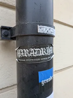 Street sticker The sticker is rectangular, predominantly black with white text, and is applied to a dark grey pipe. It shows signs of wear, with edges peeling slightly and a wrinkled surface, indicating it's a physical sticker. The main text, "HARADRIM", is rendered in a highly stylized, dripping, gothic or blackletter font, giving it a rugged, almost icy appearance. Below this, in a smaller, more traditional but still gothic-style font, is the phrase "SWEDISH BLACKENED DEATH METAL". The overall aesthetic is dark and aggressive, typical of the metal music genre it represents.