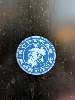 Street sticker A round blue sticker with the text 'Suptras Rostock' arranged circularly around a central image of a bearded man in a helmet, reminiscent of a Viking. The style is simple and bold.
