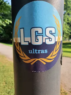 Street sticker Oval sticker with the letters LGS in the center, surrounded by a golden laurel wreath. Below the letters, the word "ultras" is written in a smaller font. The background is blue, and the laurel wreath and letters are in white and gold. The sticker is applied to a dark gray pole.