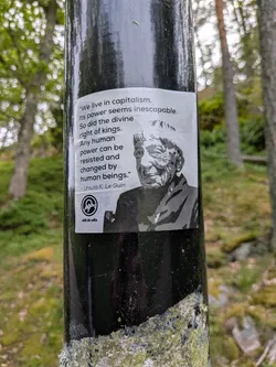 A rectangular black-and-white sticker on a pole. It displays a high-contrast portrait of Ursula K. Le Guin alongside her quote about overcoming capitalism. The bottom-left corner features the 'Allt åt alla' logo.
