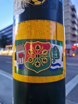 Street sticker A colorful sticker affixed to a pole. The sticker displays a crest-like emblem with various symbols, including what appears to be a star, letters, and other design elements. The colors are predominantly red, green, and gold, and the style evokes a sense of heraldry or traditional insignia.  The emblem is surrounded by what seems to be a yellow reflective background.