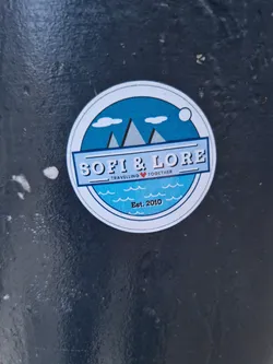 Street sticker A round sticker with a blue design featuring mountains and waves.  The text 'SOFI & LORE' is prominently displayed, along with the tagline 'Travelling ❤️ Together' and 'Est. 2010'.