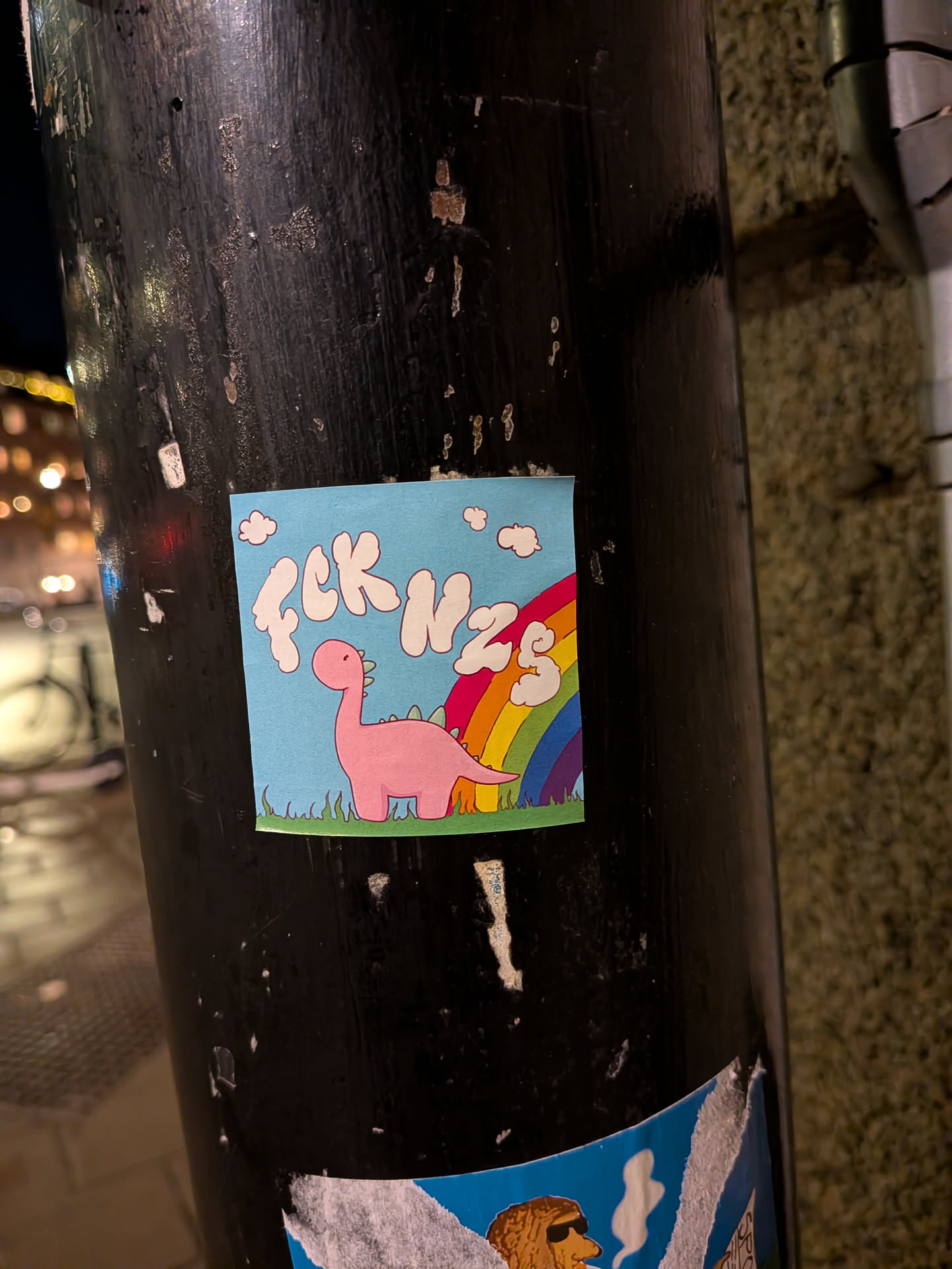 A square sticker featuring a light blue sky background with several small white cartoon clouds. In the center, a cute, pink, cartoon-style dinosaur (resembling a Brontosaurus) with small green spikes on its back stands on a strip of green grass. Behind the dinosaur, a vibrant rainbow arches upwards, displaying distinct red, orange, yellow, green, blue, indigo, and violet stripes. At the top left, the text "FCK NZS" is written in white, bubbly, cloud-like letters.