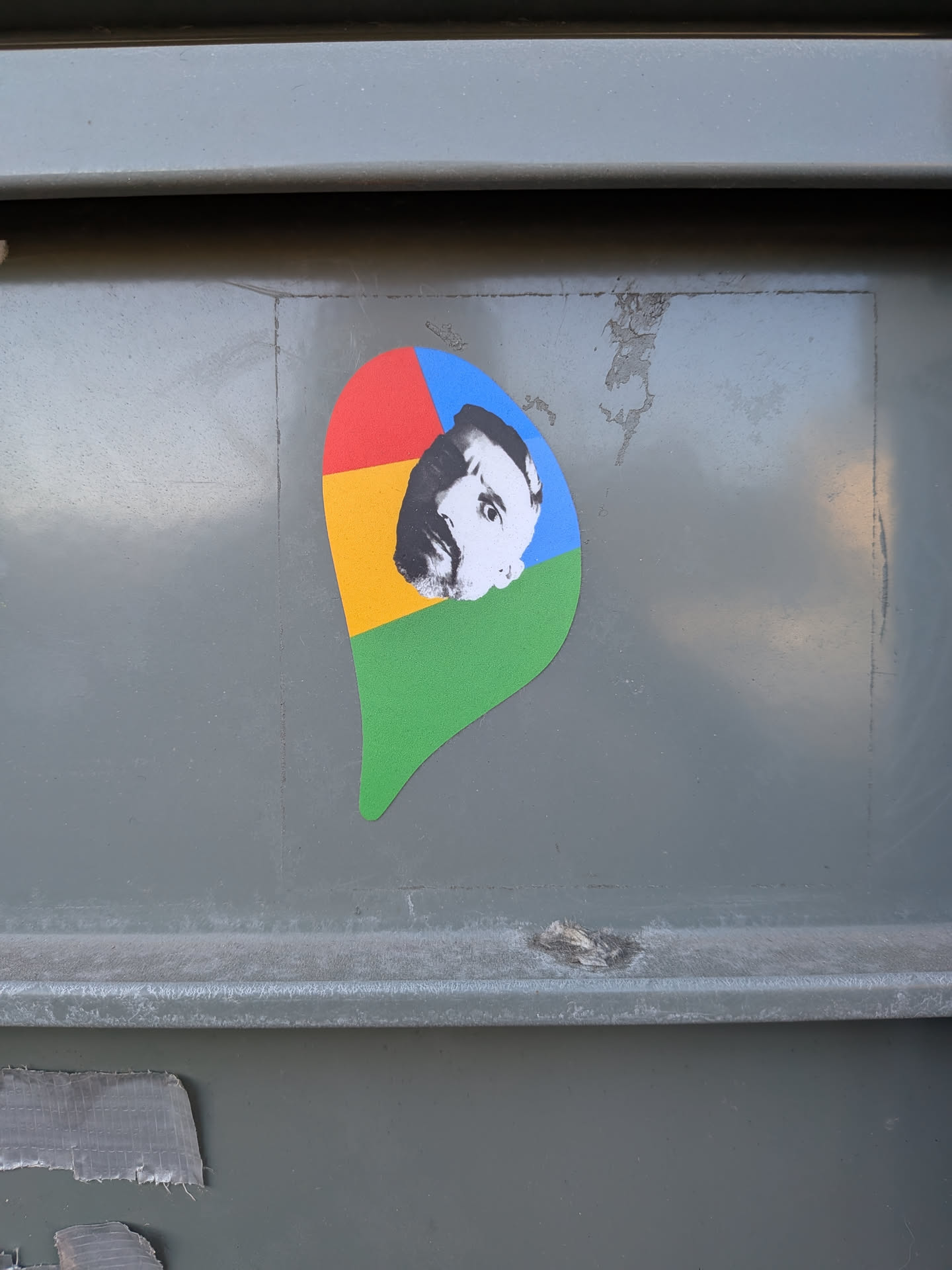 Street sticker The sticker is shaped like the iconic Google Chrome logo pin, featuring its characteristic four-color segments: red in the top-left, yellow in the bottom-left, green in the bottom-right, and blue in the top-right. Superimposed over the center of these colors is a black and white portrait of a man, strongly resembling philosopher Friedrich Nietzsche, with a prominent mustache and a serious expression. The portrait is slightly off-center, with his face primarily covering parts of the yellow, blue, and red sections. The sticker has a clean, die-cut edge and is adhered to a textured, grey metallic surface.