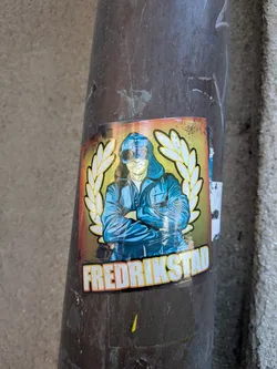 Street sticker Rectangular sticker showing a hooded figure wearing goggles with arms crossed, flanked by a yellow laurel wreath on a gradient background. Features the text 'FREDRIKSTAD' at the bottom.