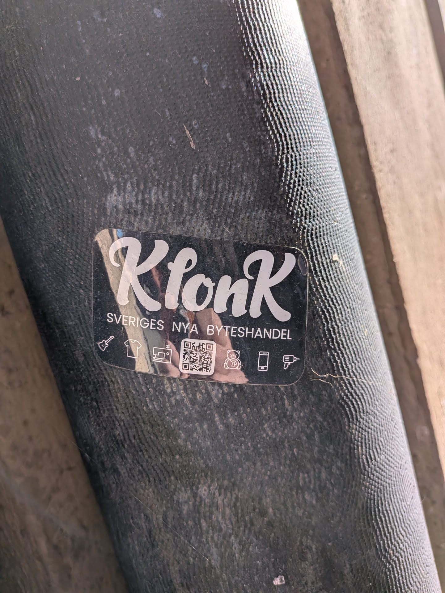 A transparent, rectangular sticker with rounded corners, featuring the brand 'KlonK' in a large, elegant white script font with a subtle shadow effect. Below the brand name, in smaller white capital letters, is the slogan 'SVERIGES NYA BYTESHANDEL' (Sweden's New Exchange/Trade). Underneath the slogan, a row of white outline icons depicts various items for trade, including a guitar, a t-shirt, a sewing machine, a teddy bear, a smartphone, and a power drill. Centered among these icons is a clear black and white QR code. The sticker is applied to a dark, textured surface, showing some reflections and minor wear.